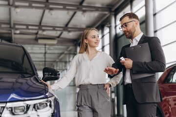 Car Sales Manager Showing Auto To Caucasian Lady Buyer Standing In Luxury Automobile Dealership Store. Buying Vehicle Concept