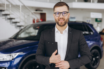 Handsome car dealership worker in suit is holding a folder and smiling while standing near the car