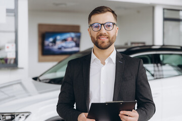 Handsome car dealership worker in suit is holding a folder and smiling while standing near the car