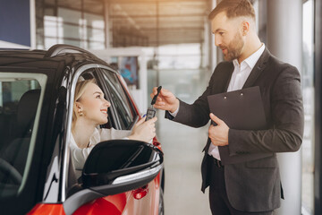 Young caucasian woman buys a car at a car dealership. Happy smiling bearded man seller gives the...