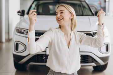 Cheerful woman standing near vehicle in dealership center and raising hands with excitement, copy...