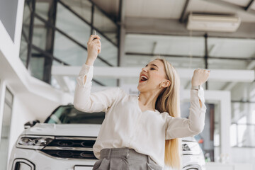 Cheerful woman standing near vehicle in dealership center and raising hands with excitement, copy space. Happy owner. Overjoyed Caucasian lady holding keys of new car after buying.