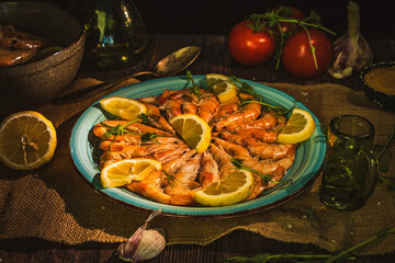 Boiled shrimp on a turquoise dish with lemon, herbs around the plate, tomatoes, garlic, sauce, a...