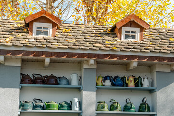 A charming outdoor shelf holds a collection of teapots beneath a roof adorned with autumn leaves....