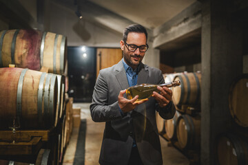 Adult man winemaker hold and check bottle of wine between barrels