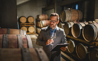 Adult man winemaker hold glass stand between the barrels in cellar