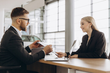 Smiling happy male manager car salesman in car dealership signing contract at table with female buyer in suit