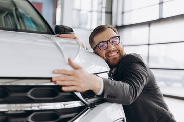 Positive young bearded caucasian man in glasses hugging his new car in dealership. Happy man...