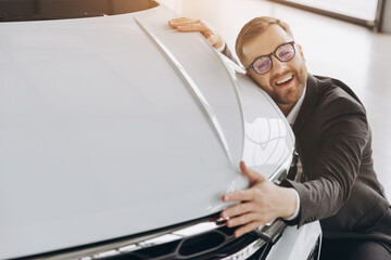 Positive young bearded caucasian man in glasses hugging his new car in dealership. Happy man...