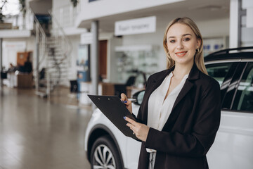 Handsome car dealership female worker in suit is holding a folder and smiling while standing near...