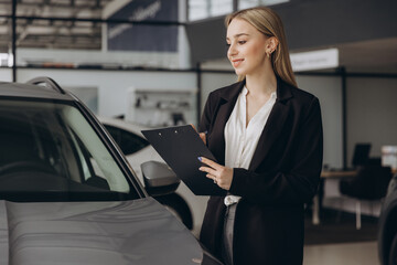 Handsome car dealership female worker in suit is holding a folder and smiling while standing near the car