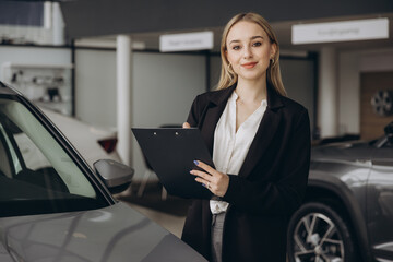 Handsome car dealership female worker in suit is holding a folder and smiling while standing near...