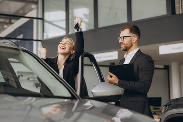 Modern happy woman raising hand up making fist near her new car in car showroom. Bearded seller...