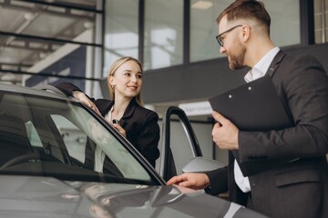 Beautiful young woman is talking to handsome car dealership worker while choosing a car in...