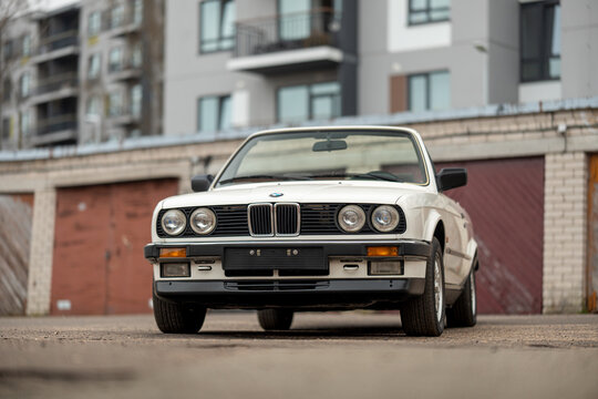Riga, Latvia - April 1, 2024: The white classic convertible BMW E30 car with opened roof. An old garages and modern building on the background.