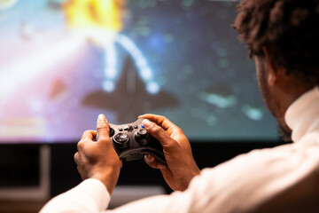 Controller held by gamer sitting in front of large projector displaying videogame. Close up shot of gamepad used by man enjoying gaming session on console connected to ultrawide smart TV