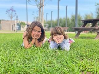  Two smiling children lying on the grass in a park, enjoying a sunny day outdoors with trees and picnic tables in the background.
