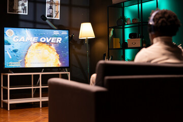 Gamer wearing headphones frustrated by seeing game over message on smart TV while playing arcade...