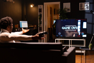 Gamer feeling gutted after losing SF videogame level, screaming at television set in anger. Angry man at home yelling, frustrated after seeing game over screen on smart TV display © DC Studio