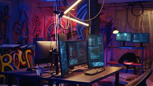 Panning shot of high tech computer system running malicious code in empty basement. PC monitors in empty neon lit criminal hideout used by hackers to commit illegal activities
