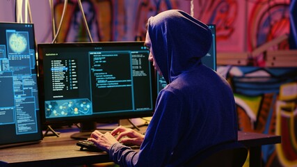 Hacker in graffiti painted hideaway base writing lines of code on computer, developing malware that...