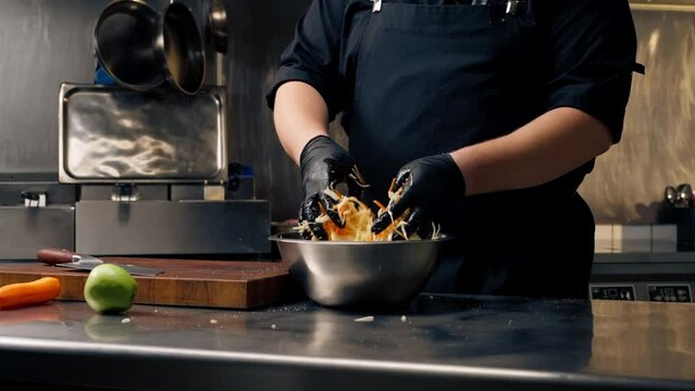 close up in professional kitchen wearing black gloves mixes chopped vegetables in an iron bowl