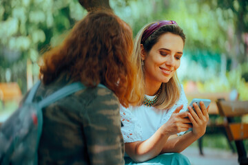 Women Talking Holding Smartphone Checking Social Media. Friends talking in the park looking at the...