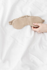 Beige eye mask for sleep in woman hand on white bedclothes, minimal lifestyle aesthetic photo. Top...