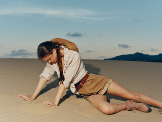 Young woman in shorts and hat crawling on top of sandy dune under clear sky in desert landscape