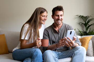 Smiling young caucasian couple relaxing at home using phone together looking social media.