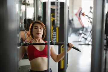 Sport determined woman in a red sports bra and black leggings uses a lat pulldown machine in a modern gym. Her focused expression and fit physique highlight her commitment to strength training.