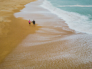 A couple walks on the beach of Nazaré during the waves on a spring day, drone view
