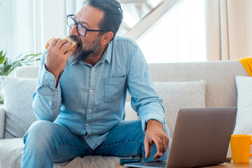 One man in casual clothes using laptop at home sitting on sofa and eating sandwich alone. People with food and technology. Indoor leisure activity. Computer and nutrition. Surfing the net connection - Powered by Adobe