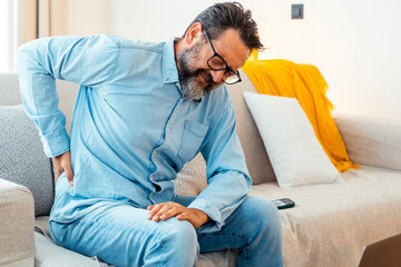 Fototapeta premium One man massaging his lower back after using laptop sitting on sofa for bad posture sitting. People unhealthy habits workplace at home. Mature male massage his body with painful expression on face