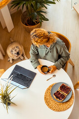 Top vertical view of woman in morning lifestyle having breakfast while use laptop to check emails messages and notifications to start the working day. Small puppy on the floor. Remote worker lifestyle