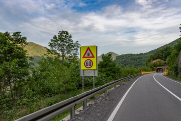 triangular road sign indicating a warning, with a speed limit sign of 60 km/h below it. This is situated on a mountain road with a tunnel entrance visible in the background.