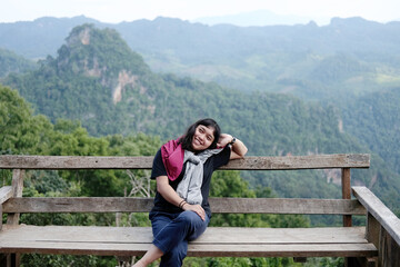 Naklejka premium Asian woman sitting and relaxing on wooden bench on terrace for freedom journey with valley mountain and foggy in natural and travel destination in tropical rain forest