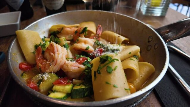 Putting grated parmesan cheese on a plate of paccheri al Profumo di Mare. The dish features al dente pasta, succulent shrimp, and a medley of fresh vegetables, all garnished with aromatic herbs for an