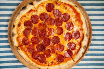 Top view hot pepperoni pizza with sausage cut into portions on a striped tablecloth in a pizzeria on the table