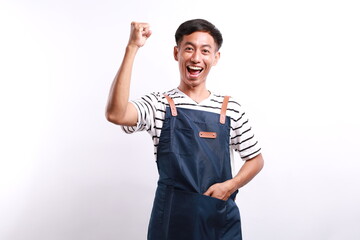 Cheerful asian seller making fist pump, rejoicing and triumphing, standing in blue apron against...