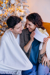 Joyful couple sits close together by beautifully decorated Christmas tree. The woman is holding...