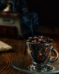 Coffee beans of varying degrees of roasting in a transparent coffee cup on a wooden table with manual coffe grinder and smoke in the background