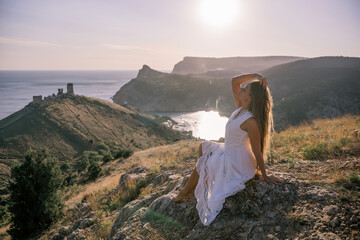 A woman in a white dress is sitting on a rock overlooking a body of water. She is enjoying the view...