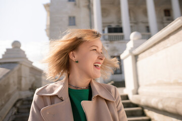 A woman with long hair is standing on a set of stairs. She is wearing a green shirt and a tan coat.