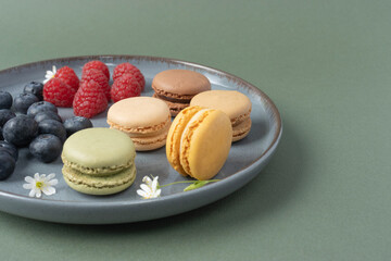 Macarons in a plate on a green background. Berries and pastries close-up with copy space. Strawberries, raspberries and blueberries are decorated with white flowers. French breakfast. Tea party