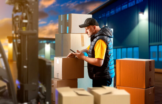 Man is logistician. Storekeeper stands among boxes. Loader with parcels near night factory. Logistician checks data on parcels. Warehouseman accepts delivery. Man prepares goods for shipment - Powered by Adobe