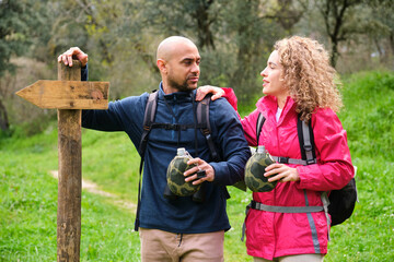 A man and a woman are resting standing next to a sign with canteens in a forest. They are carrying...
