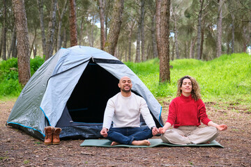 A man and woman are sitting by a tent in the woods meditating. They are both in a relaxed state,...