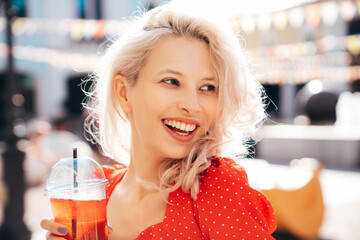 Young beautiful smiling hipster woman in trendy summer clothes. Carefree model posing in the street. Holding and drinking fresh vegetable cocktail smoothie drink in plastic cup with straw