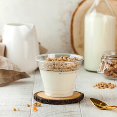 Natural yogurt topped with crunchy muesli in a clear cup with a milk jug and a jar of muesli in the background